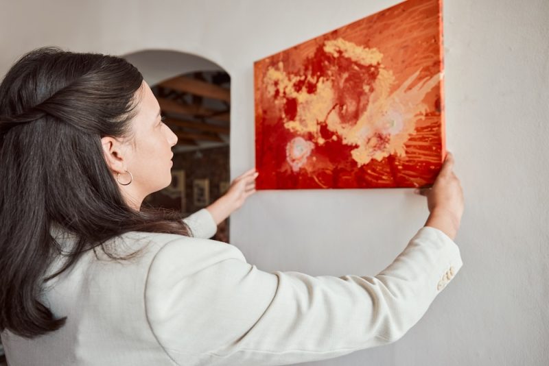 A woman hangs up a canvas painting in her home. The painting is abstract with red and orange splashes.