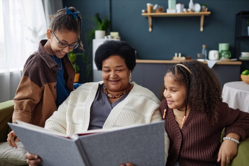 An older black woman sits with her two grandchildren on a couch. All are smiling and looking at a photo album.