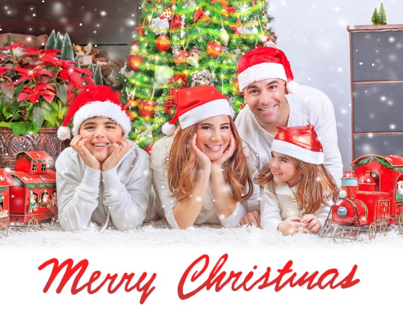 A family of four lies down and poses for a Christmas card. There is a Christmas tree behind them, and all are smiling and wearing Santa hats.